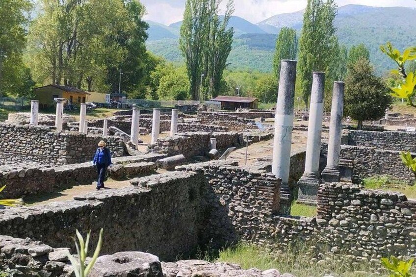 Ruinas antiguas con muros y columnas de piedra, una persona de pie, y un paisaje montañoso al fondo. 4