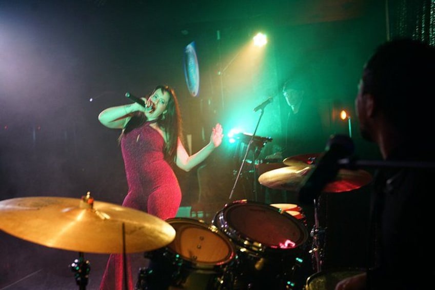 A person singing on stage with a microphone, a drum set, and a keyboard in the background. 2
