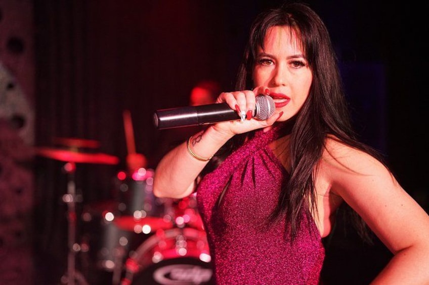 A person holding a microphone, wearing a sleeveless top, with a drum set in the background. 2
