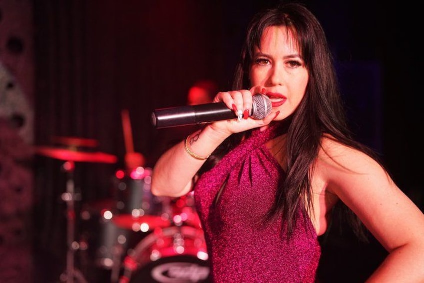 A person holding a microphone, wearing a sleeveless top, with a drum set in the background. 4