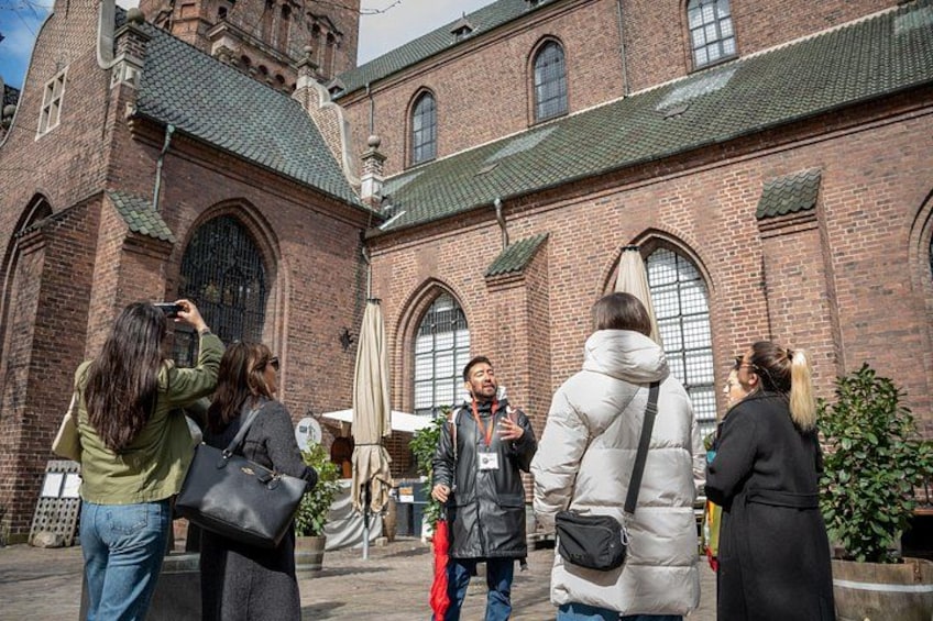 A group of people standing in front of a historic brick building with arched windows and a tower. 2