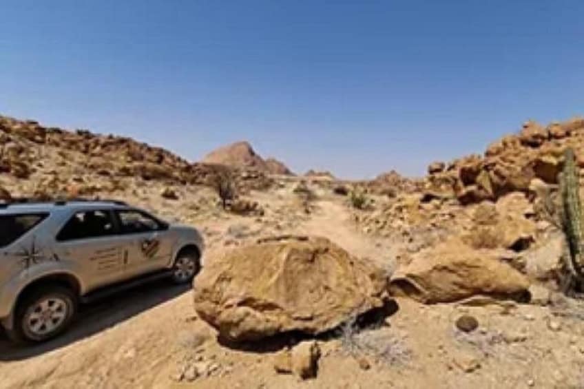 Un paysage désertique avec un véhicule stationné sur un terrain rocheux, de gros blocs de pierre, une végétation clairsemée et un ciel dégagé. 5