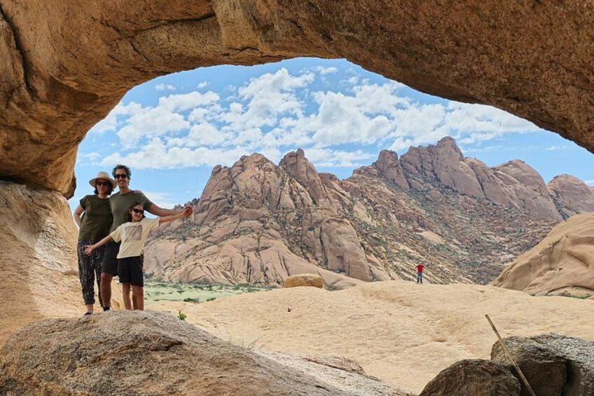 Une famille se tient devant une arche naturelle formée par des roches, avec un paysage montagneux en arrière-plan. 3