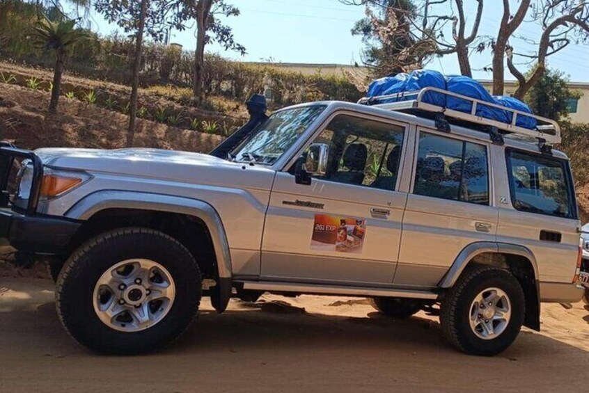 A silver SUV with a roof rack and blue tarps on the roof, parked on a dirt road with sparse vegetation and trees in the background. 1