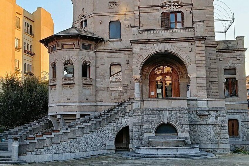 A historic stone building with a central arched doorway, a staircase leading to the entrance, and a small fountain in front. 5