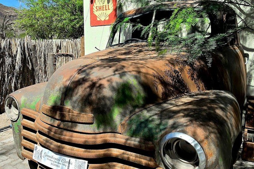 Un coche antiguo con carrocería oxidada y una estación de servicio Shell al fondo. 5