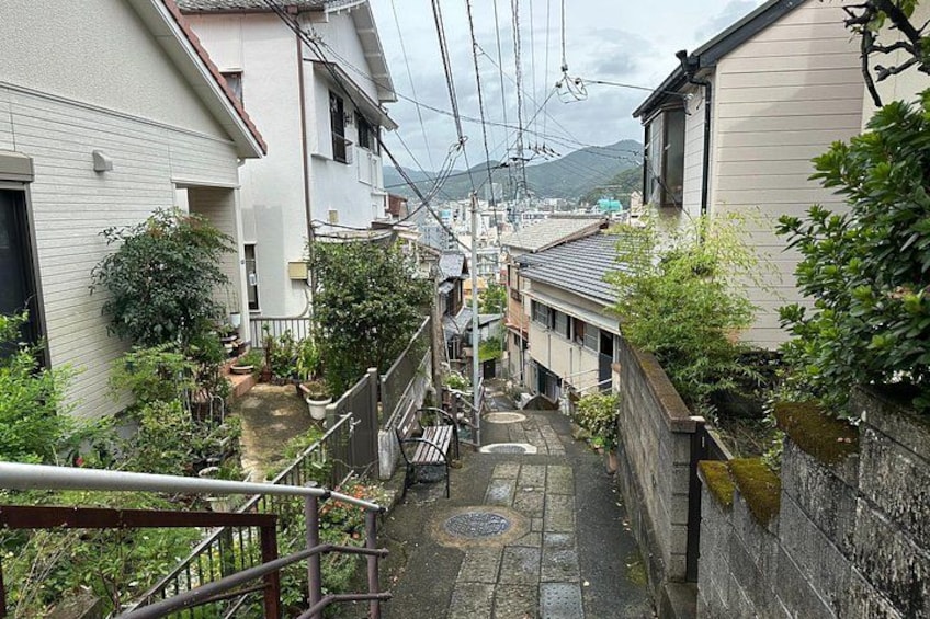 Ein schmaler Gang zwischen traditionellen japanischen Häusern mit einer kleinen Gartenanlage, einem Metallgeländer und einem entfernten Berg. 2