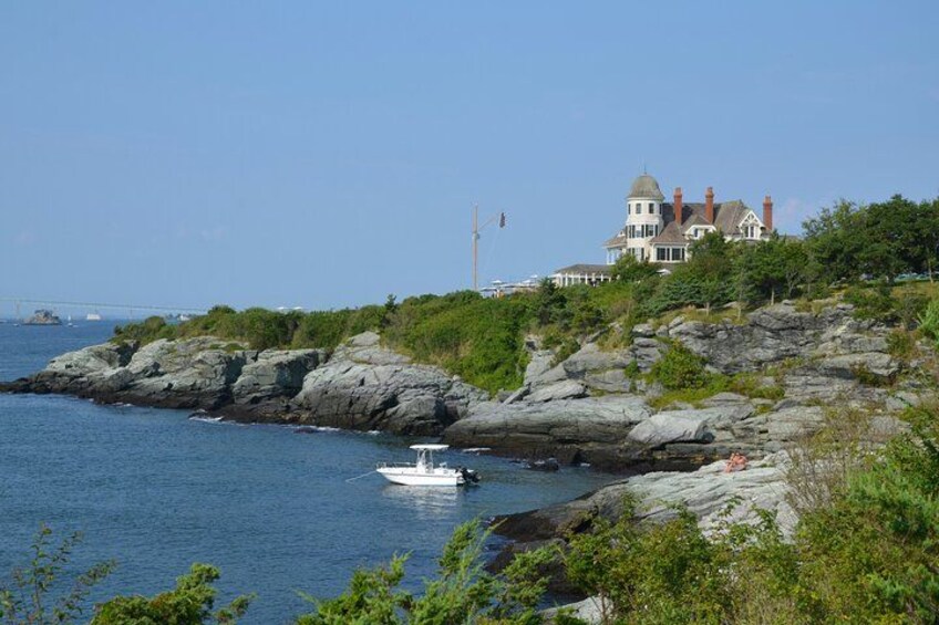 A coastal landscape with a large house on a rocky outcrop, a boat in the water, and a bridge in the distance. 1