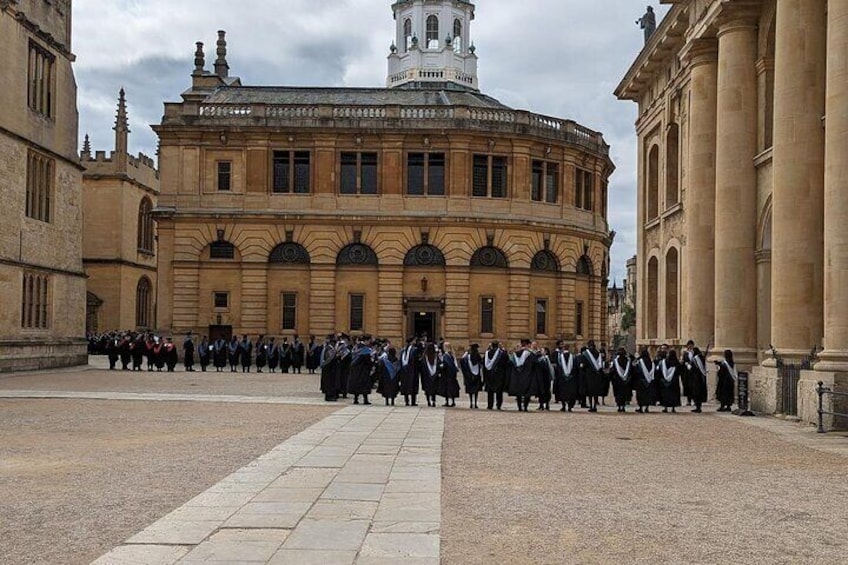 A group of people in academic regalia gathered in front of a historic building with a round central section and a clock tower. 4