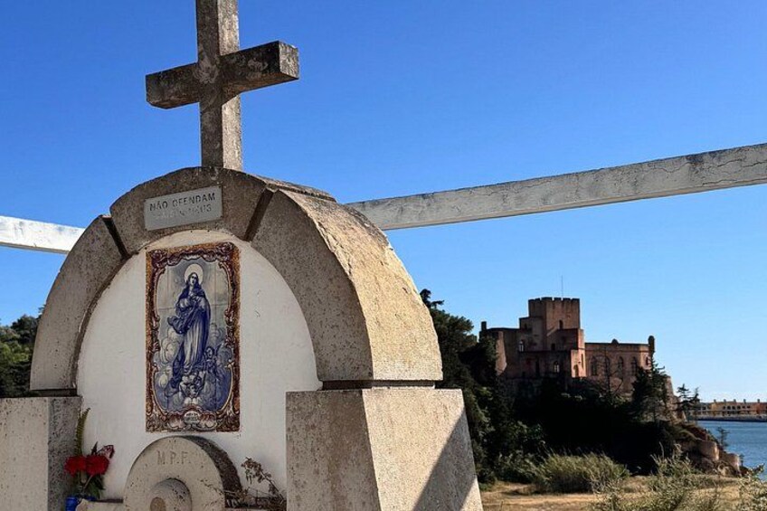 Un monumento histórico con una cruz, una placa conmemorativa y una estatua de una mujer. En el fondo, se observa un castillo junto al agua. 2