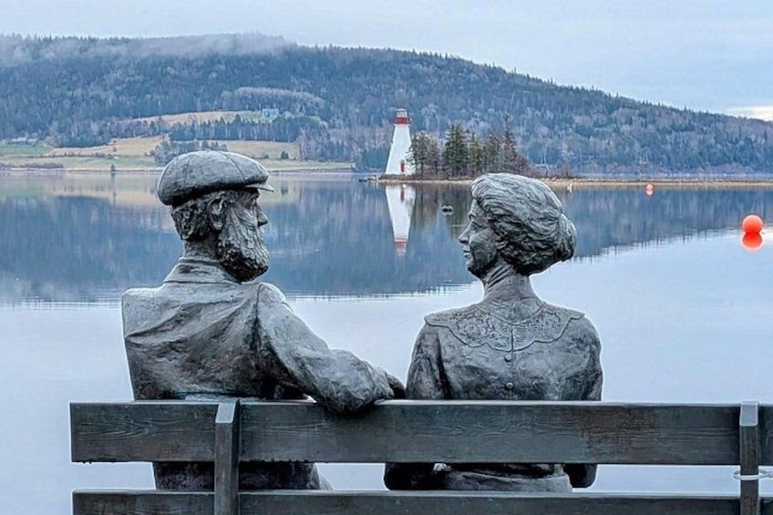 A statue of a man and woman sitting on a bench by a lake with a lighthouse in the background. 2
