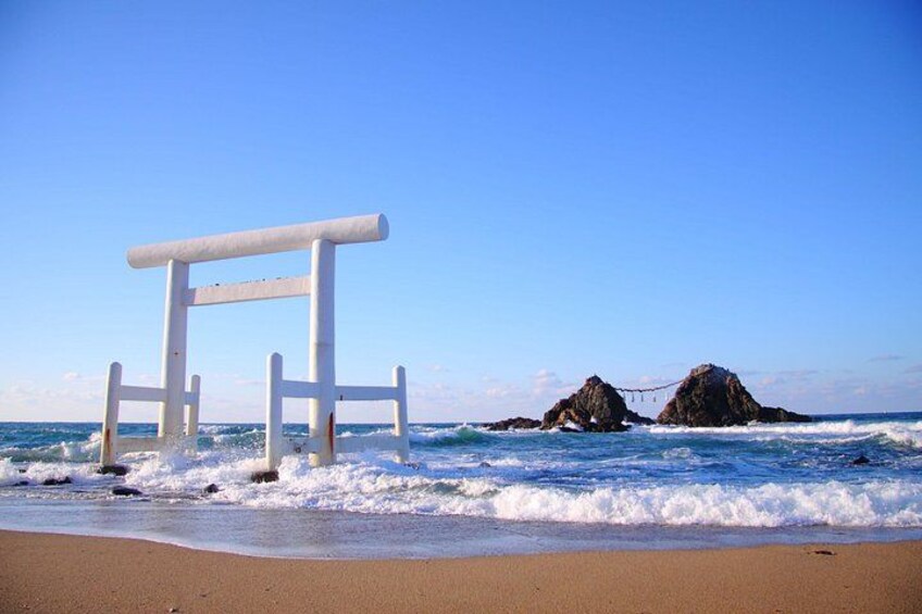 A white structure on a beach with waves crashing around it and two rocky formations in the background. 1