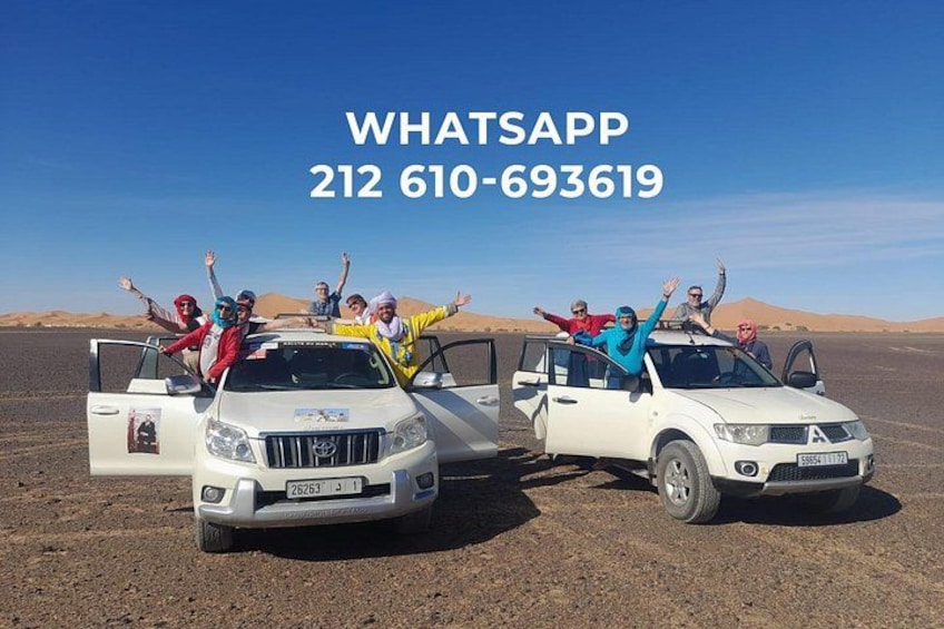 Two white SUVs with passengers raising their hands in the desert. 1