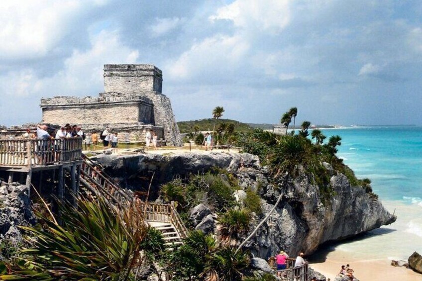 A coastal archaeological site with a large stone structure, a wooden viewing platform, and a beach. 3