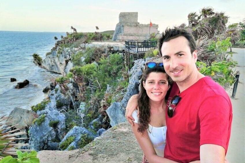 A couple on a rocky coastal path with a view of the sea and a historical structure in the background. 1