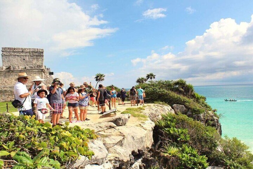 A group of people standing on a rocky cliff overlooking turquoise waters, with a historical stone structure in the background. 4