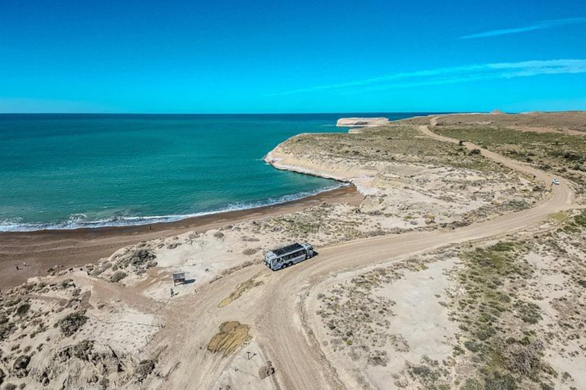 A coastal landscape with a winding dirt road, a parked vehicle, and a distant building by the sea. 3