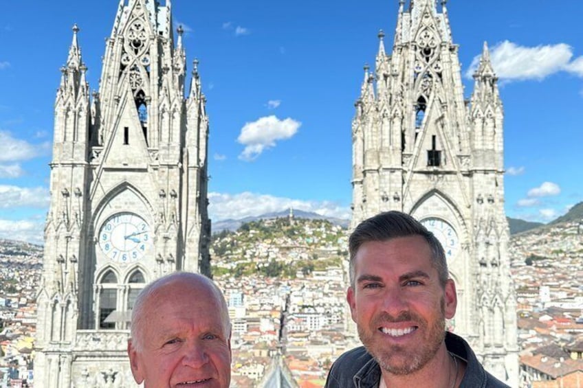 Two individuals in front of a Gothic cathedral with a clock face, set against a cityscape and mountains. 3