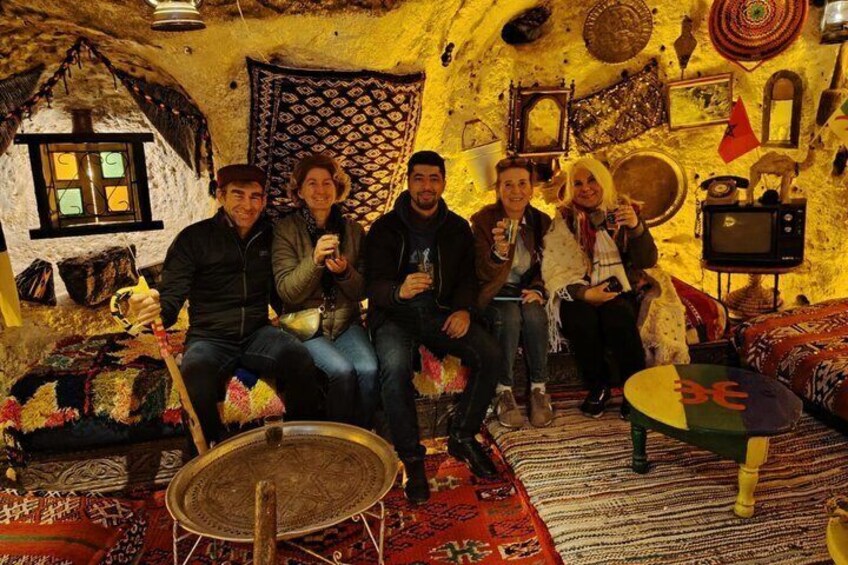 Un gruppo di persone sedute in una stanza con decorazioni tradizionali, tra cui un tappeto a motivi, una piccola struttura simile a una capanna e un tavolo rotondo. 1