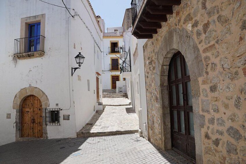 A narrow cobblestone street flanked by buildings with white walls and stone facades, wooden doors, and balconies. 2