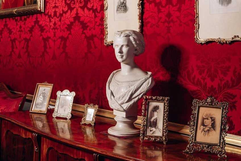 A bust sculpture on a wooden table with ornate red wallpaper and framed pictures. 2