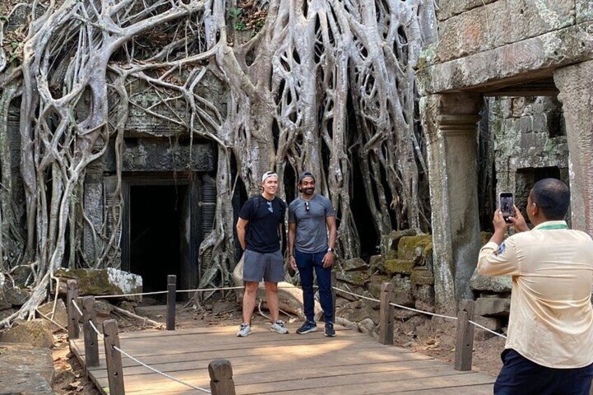 Two people standing on a wooden bridge with roots and stone structures in the background. 2