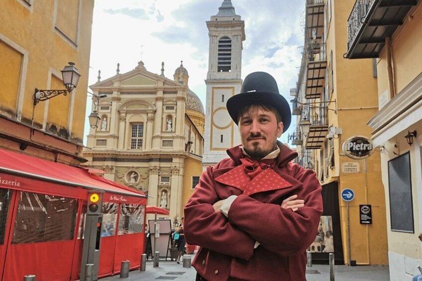 En person står på en gade i en by med historiske bygninger, et klokketårn og et rødt markise med ordet 'Pizzeria'. 2
