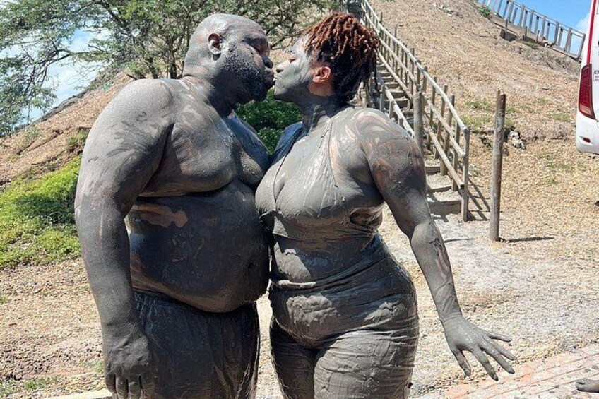 Two people covered in mud, standing on a dirt path with a wooden staircase and trees in the background. 1