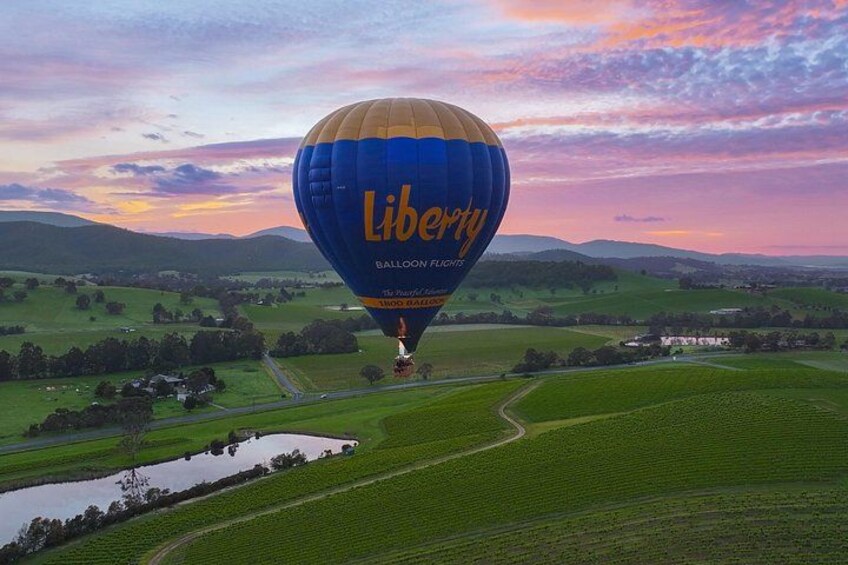 A hot air balloon with 'Liberty Balloon Flight' written on it, flying over a green landscape with fields and a small body of water. 2