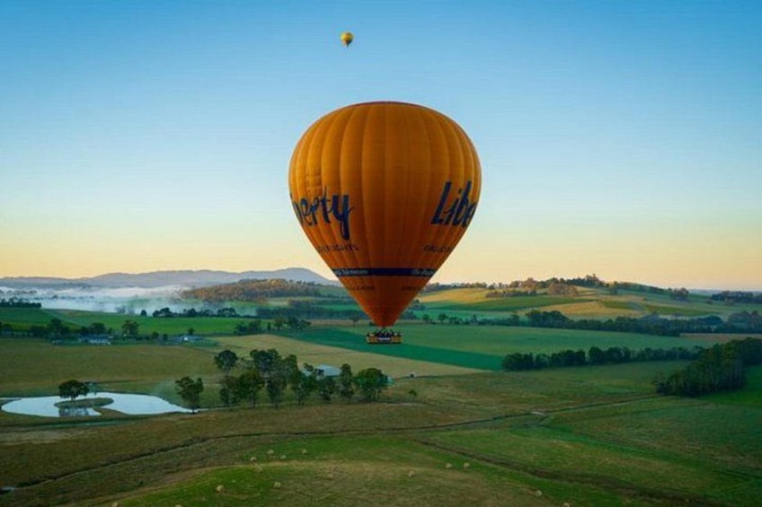 A hot air balloon with 'Lita' written on it, flying over a rural landscape with fields and a small body of water. 1