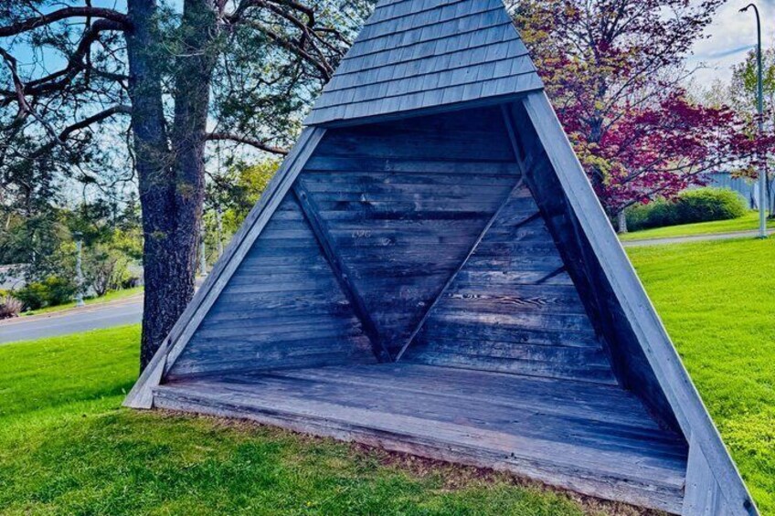 Une structure triangulaire en bois, dotée d’un toit incliné, entourée d’herbe et d’arbres. 4