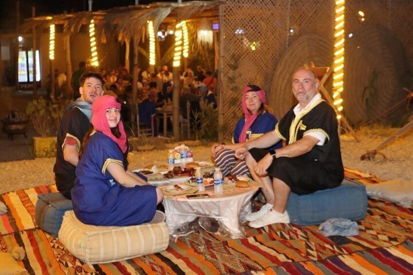 A group of people sitting on a colorful blanket outdoors at night, with string lights in the background. 5