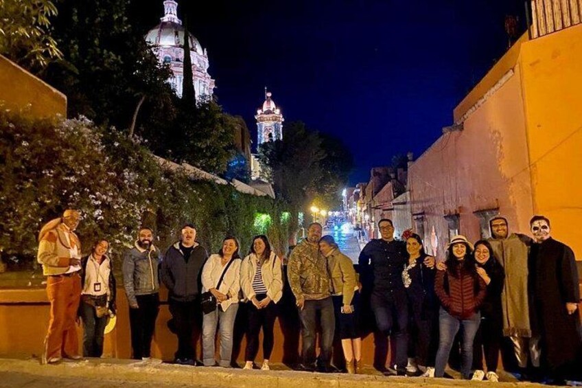 A group of people standing on a street at night with illuminated buildings in the background. 4