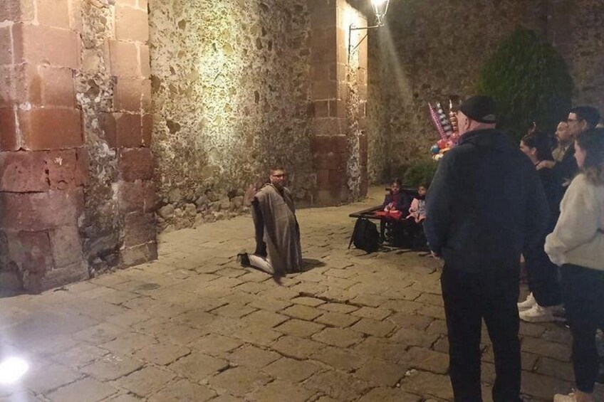 A person in traditional attire standing in a stone-walled courtyard with a crowd of people around. 1