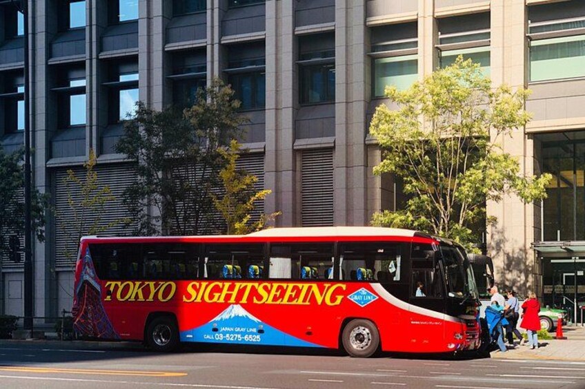 Een rode en blauwe toeristenbus met 'TOKYO SIGHTSEEING' op de zijkant, geparkeerd voor een modern gebouw met grote ramen. 2