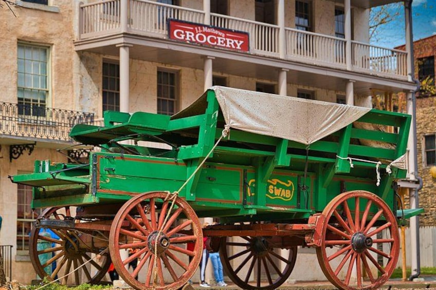 A green horse-drawn carriage with red wheels parked in front of a building with a sign that reads 'GROCERY'. 2