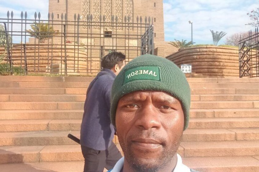 A person in a green beanie with a logo, standing in front of a building with steps and a fence. 4