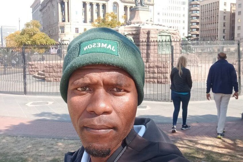 A person wearing a green beanie with 'LAMBETH' on it, standing in a park with a statue and buildings in the background. 2