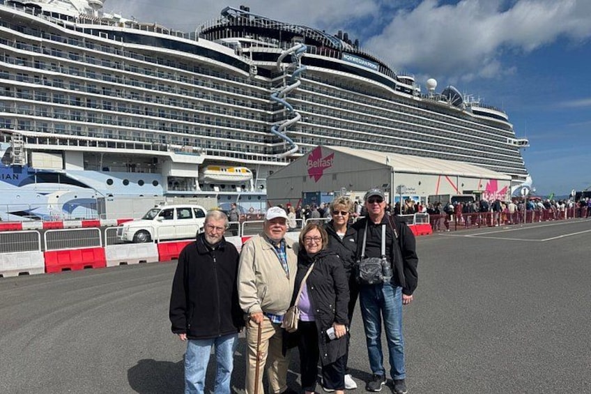 Un grand navire de croisière, doté de plusieurs ponts et arborant le logo « Belfast », est amarré dans un port où une foule de personnes s’est rassemblée, et une barrière rouge est visible au premier plan. 2