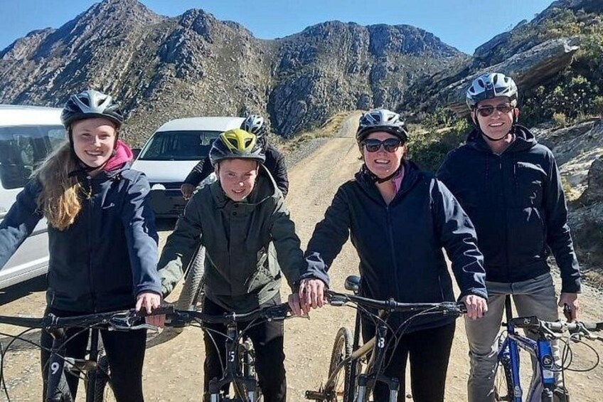 Fire personer på cykler, en hvid bil og et bjergrigt landskab. 2