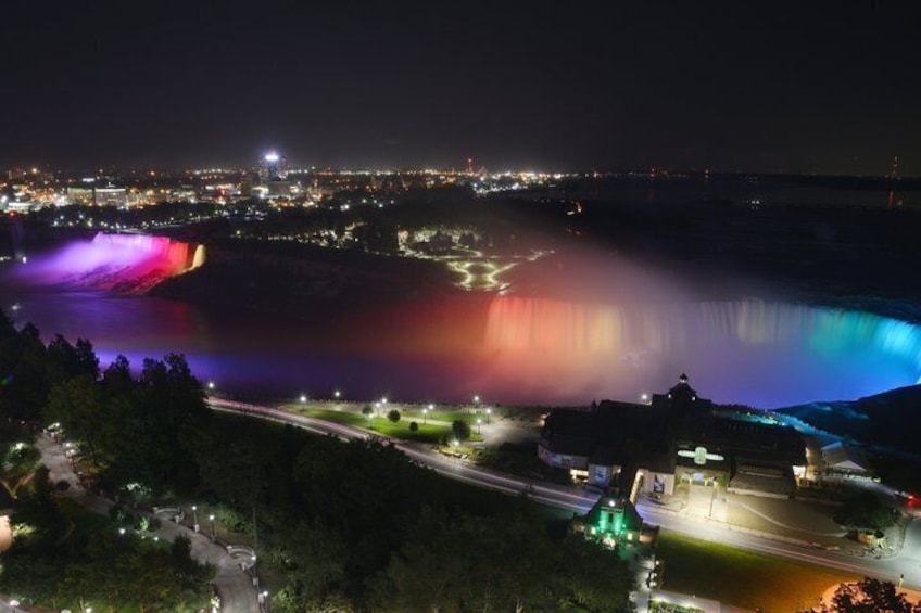 A nighttime view of a city with a waterfall illuminated by colorful lights, a lit-up bridge, and a road with vehicles. 1