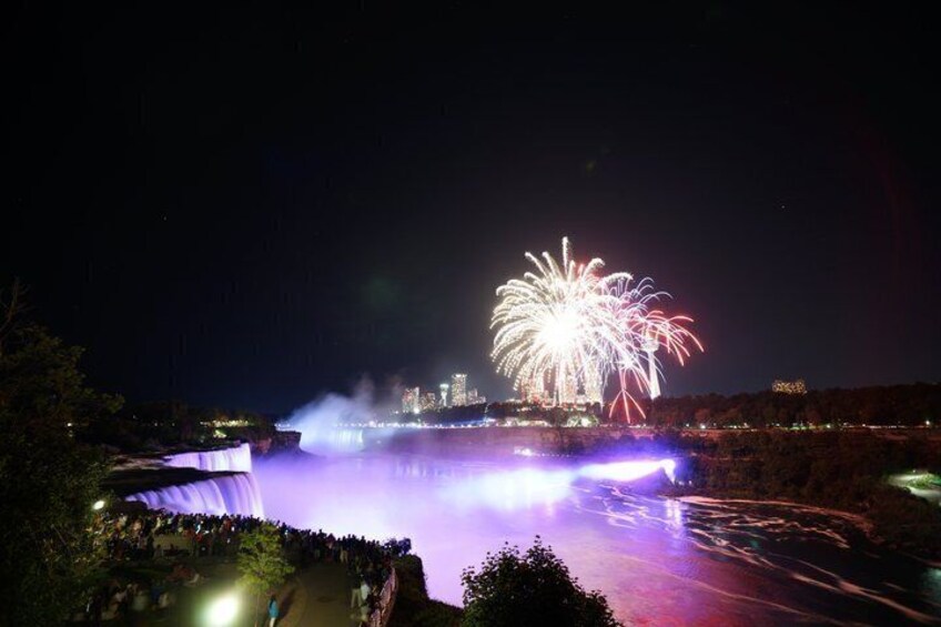 A waterfall illuminated by lights, a crowd of people, and fireworks in the night sky. 4