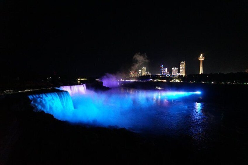 A nighttime view of a waterfall with illuminated blue and purple lights, a city skyline in the background, and a prominent tower. 3