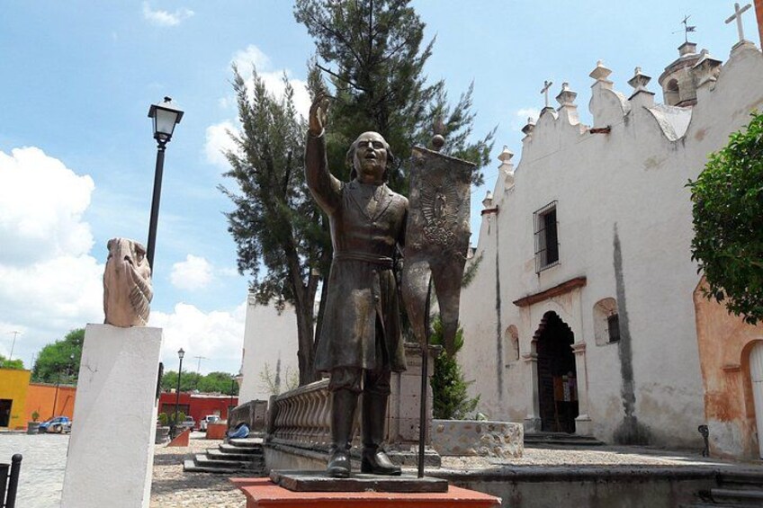Una estatua de un hombre con los brazos levantados, un edificio de paredes blancas y detalles rojos, y un farol. 1