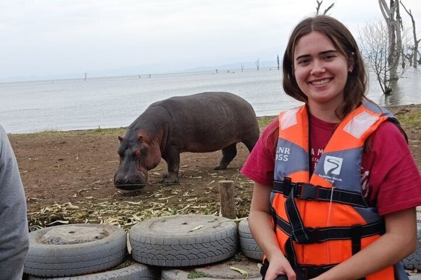 Una persona con un giubbotto arancione in piedi accanto a un ippopotamo, in prossimità di un corpo d'acqua. Si vedono pneumatici e un albero senza foglie sullo sfondo. 2