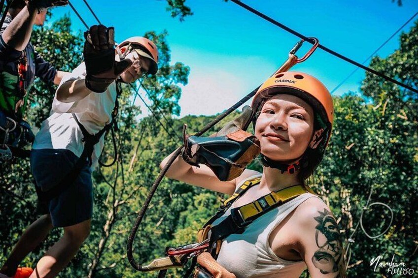 Two people engaged in a zip-lining activity, wearing helmets and harnesses, with trees and a clear sky in the background. 1