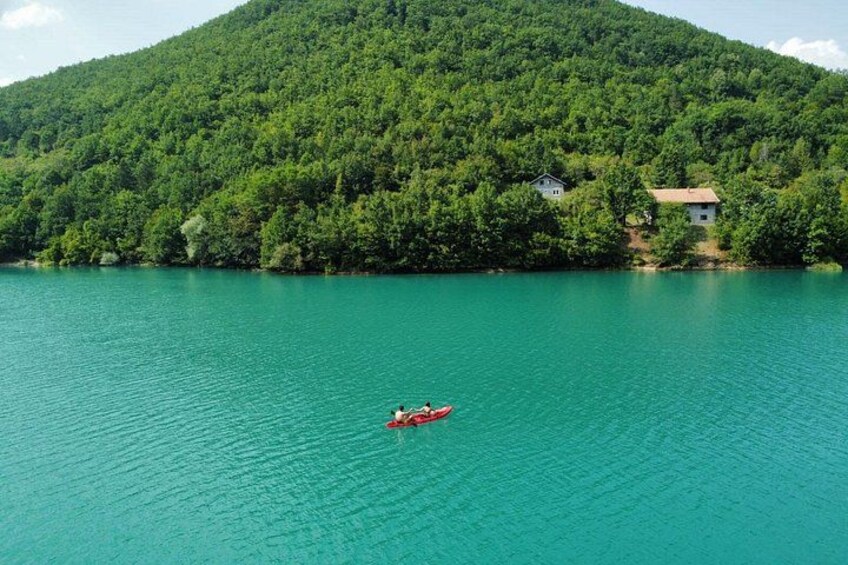 Un lago tranquilo con un kayak rojo, rodeado de un denso bosque verde y una colina con una casa. 1