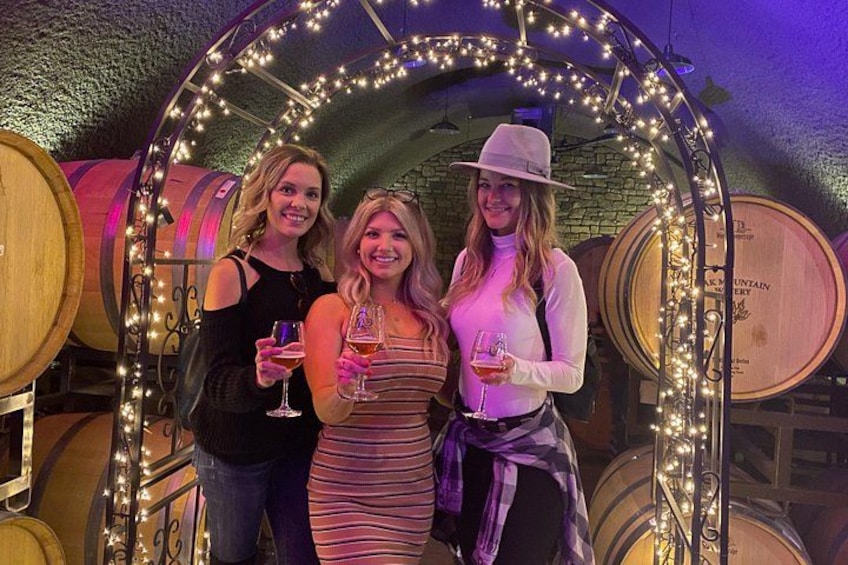 Three people standing in front of a decorative archway with string lights, surrounded by wooden barrels. 1