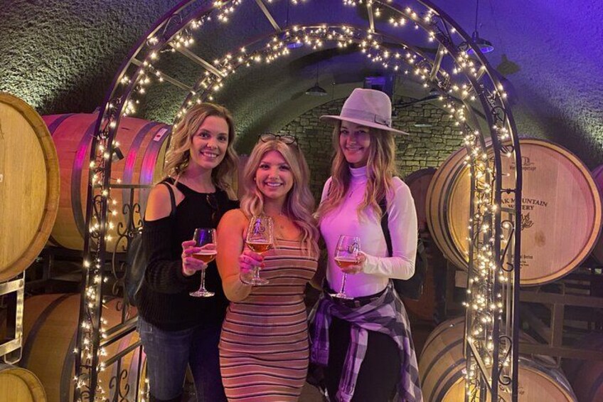Three people standing in a wine cellar with barrels and string lights. 1