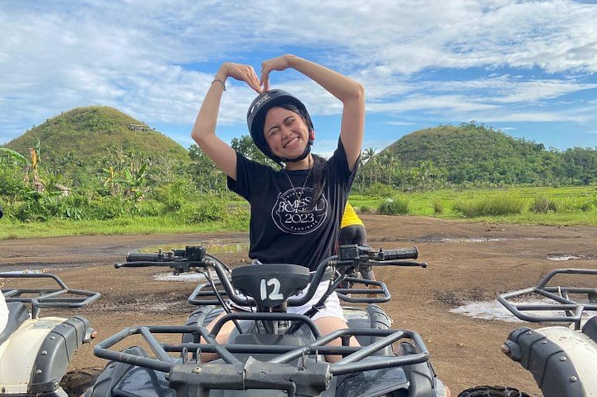 A person on an ATV with a helmet, surrounded by other ATVs, in a rural setting with hills and greenery. 5
